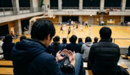 体育館の観客席で予備バッテリーを手のひらに載せて確認する撮影者の後ろ姿