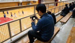体育館の2階席のベンチに座って試合を撮影する撮影者の後ろ姿